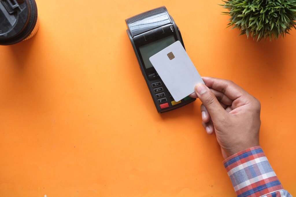 A man holds a credit card over a card scanner on an aesthetic orange background that could be noted by Spring Oaks Capital Collections